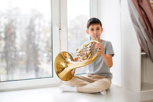 A child playing the french horn sitting with his legs crossed near a window